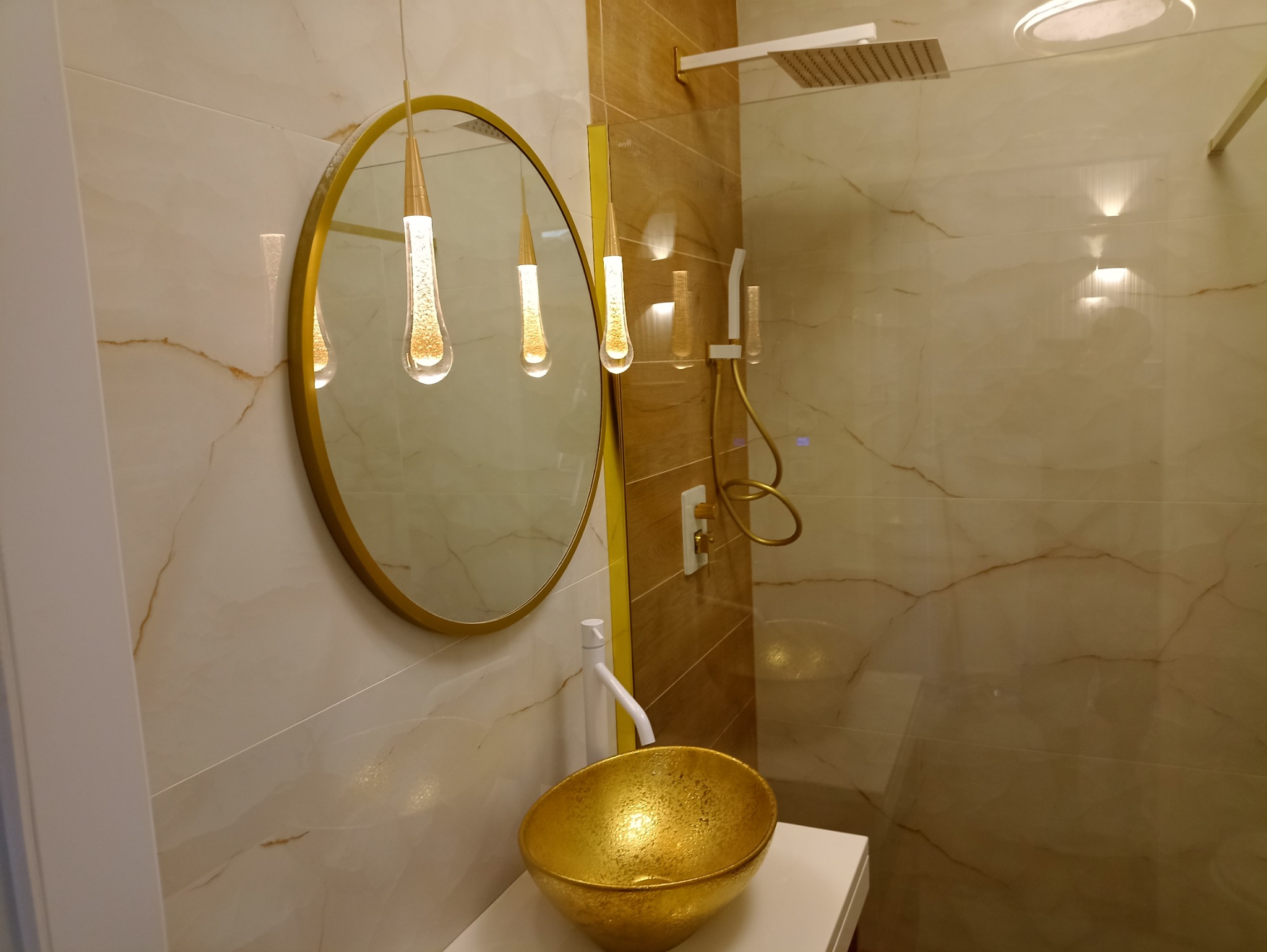 Wykończona łazienka z marmurowymi płytkami, złotą umywalką nablatową i okrągłym lustrem z podwieszonymi lampami w kształcie kropli.