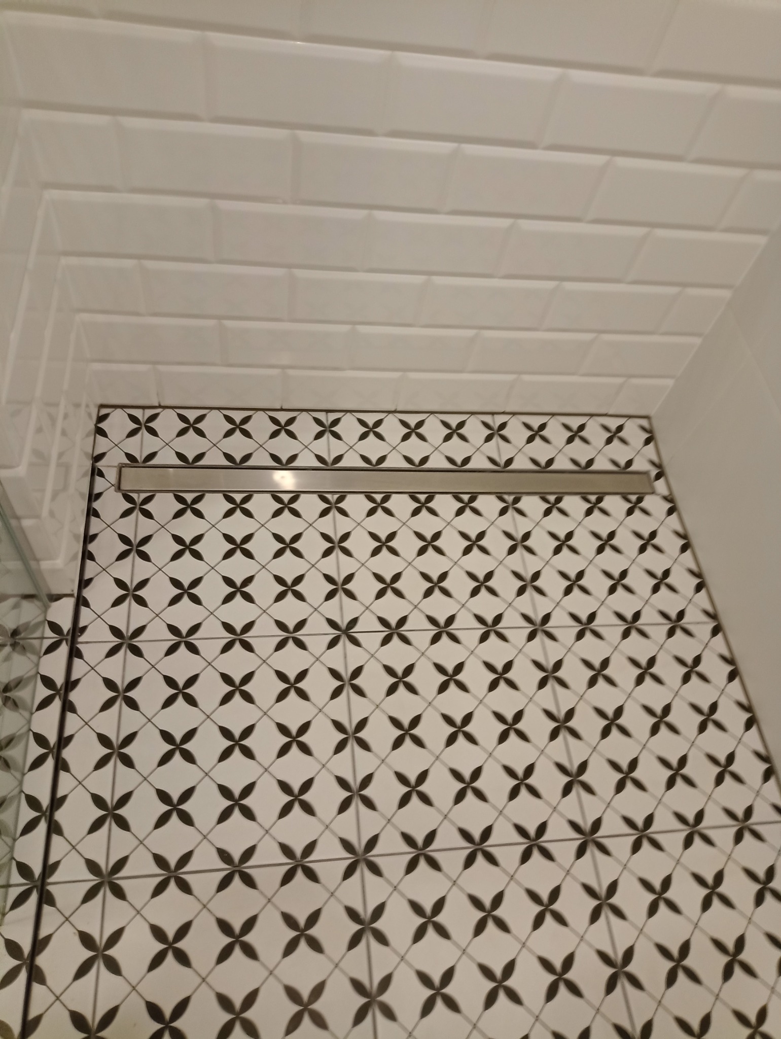 Wykończona podłoga prysznica z czarno-białą wzorzystą glazurą i odpływem liniowym oraz białe kafelki na ścianie.