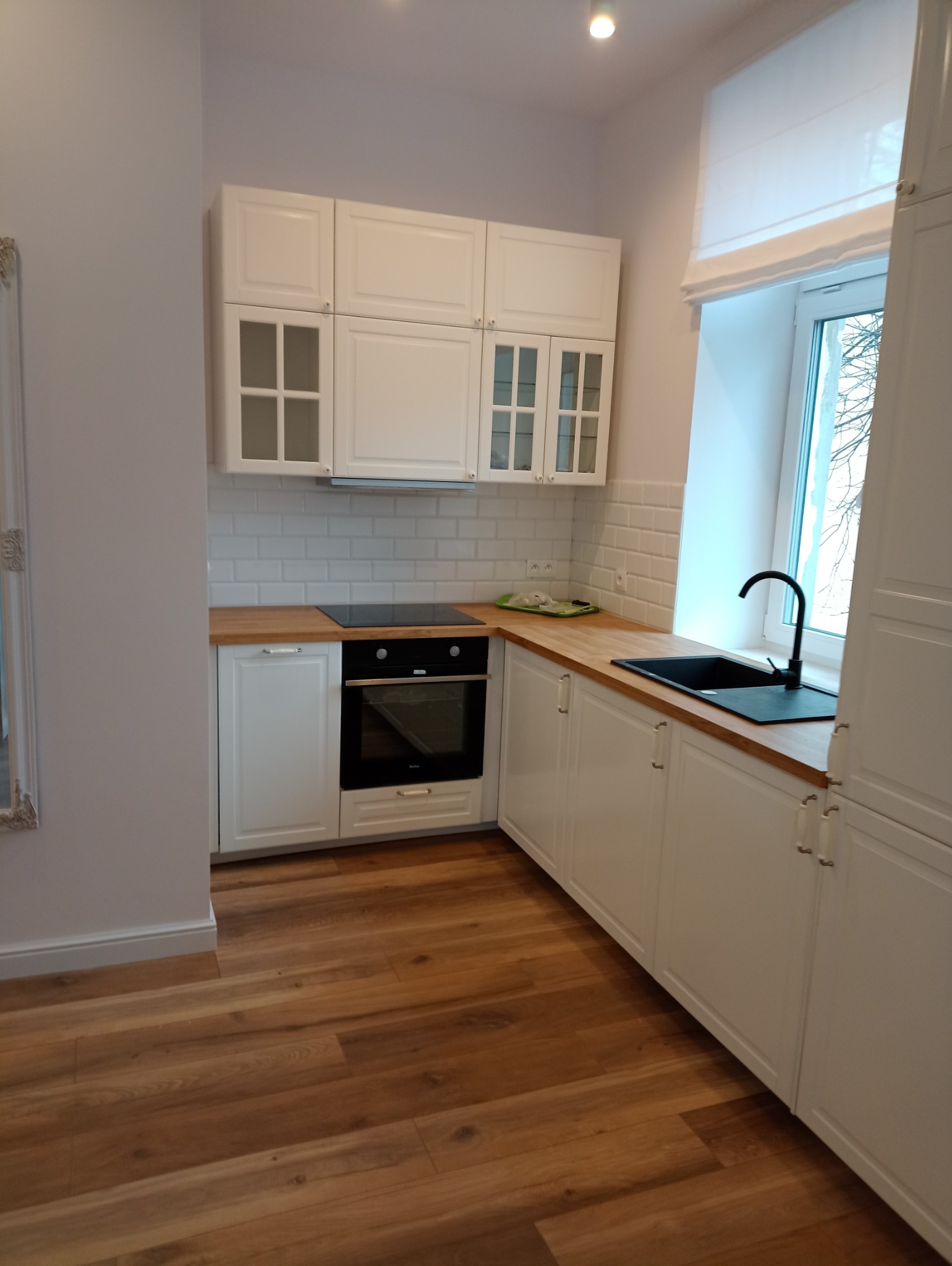 Nowoczesna, jasna kuchnia z białymi szafkami, drewnianym blatem i parkietem. Czarna bateria i zlew kontrastują z bielą, a subtelne oświetlenie podkreśla minimalistyczny styl wnętrza.