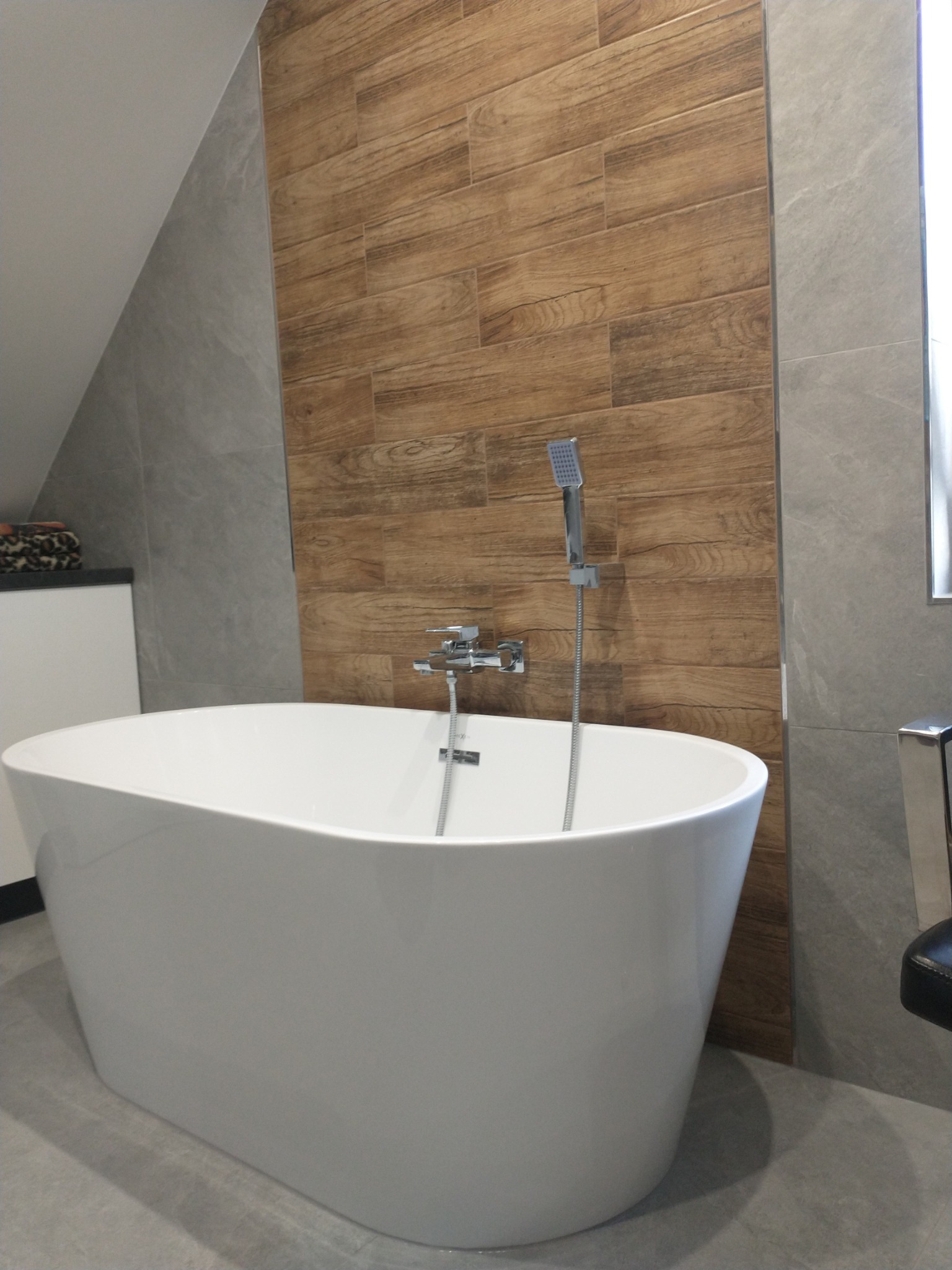 Wolnostojąca biała wanna owalna z chromowaną baterią i słuchawką prysznicową na tle ściany z płytek imitujących drewno i szarych płytek imitujących kamień.