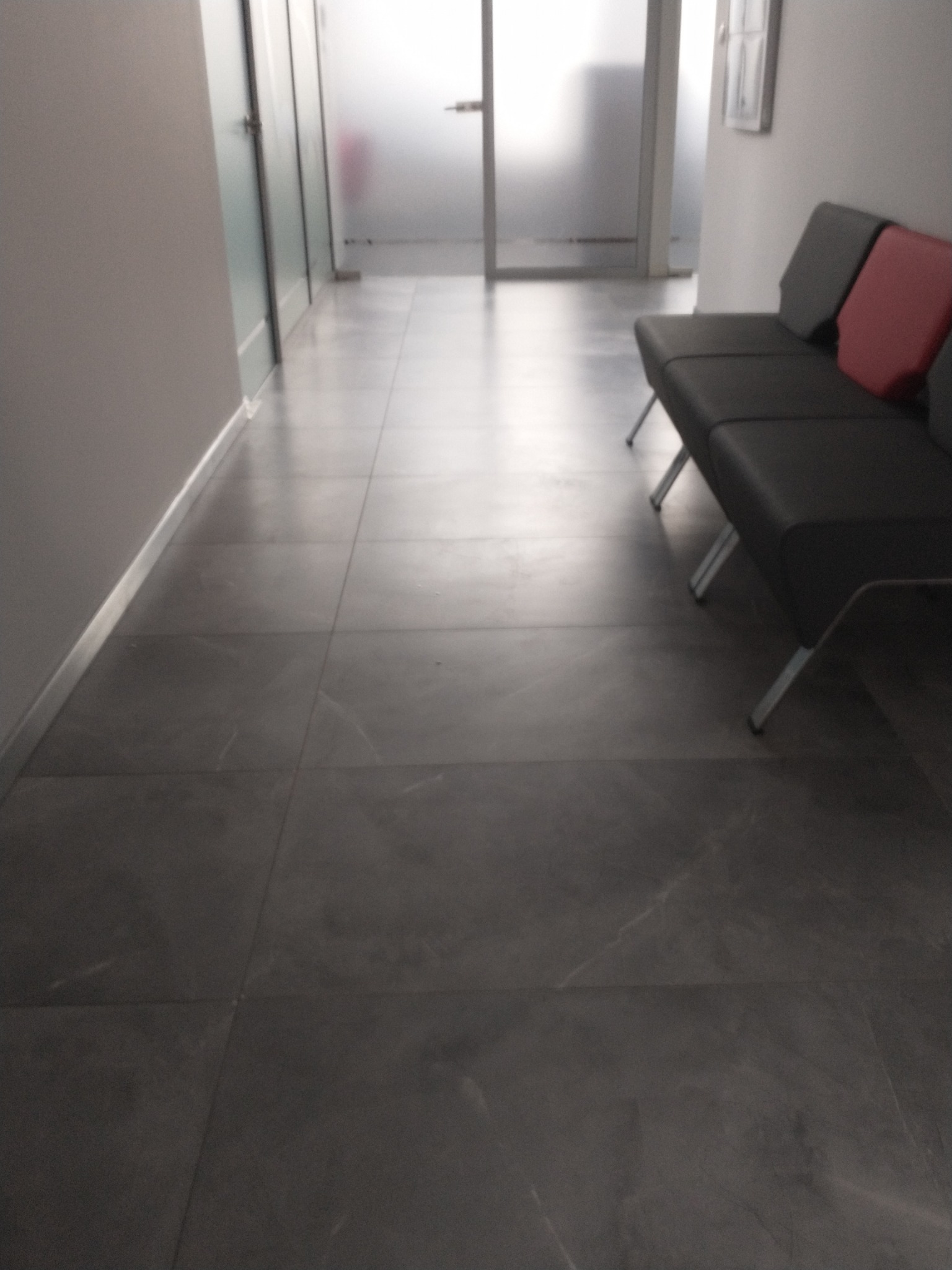 Wyłożona szarymi płytkami podłoga w korytarzu biurowym z sofą i przeszklonymi drzwiami w tle.