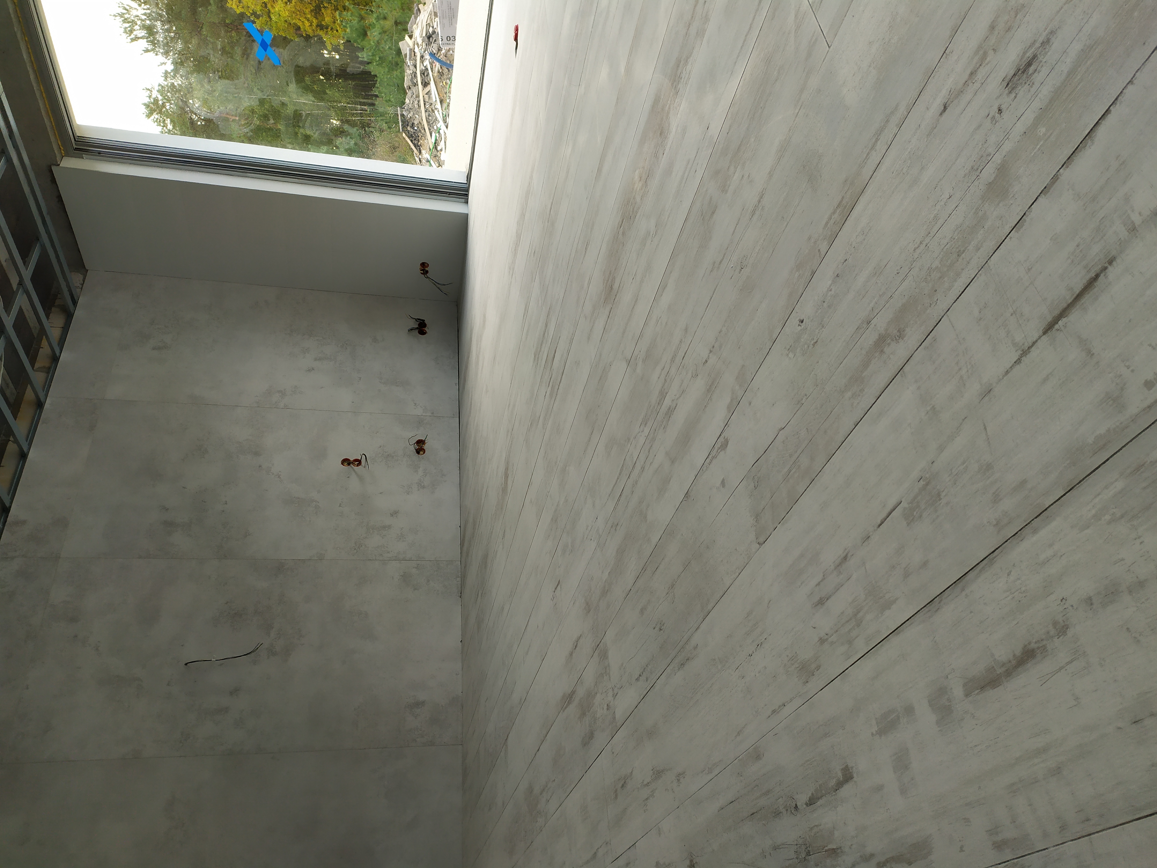 Ściana z szarych płytek imitujących beton oraz podłoga z paneli w odcieniach bieli i szarości. Widoczne wystające kable elektryczne oraz fragment okna z widokiem na zewnątrz.