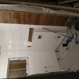 Wnętrze łazienki w trakcie remontu: białe płytki ścienne z drewnianymi akcentami, szare płytki podłogowe, widoczne narzędzia i akcesoria budowlane.