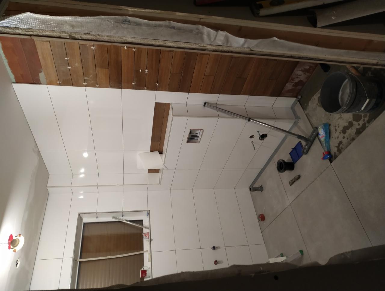 Wnętrze łazienki w trakcie remontu: białe płytki ścienne z drewnianymi akcentami, szare płytki podłogowe, widoczne narzędzia i akcesoria budowlane.