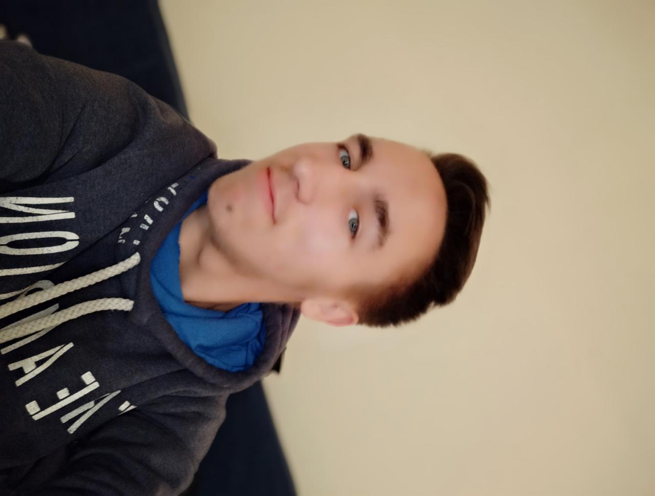 Portret młodego mężczyzny w szarej bluzie z kapturem i niebieską koszulką, z krótkimi, ciemnymi włosami i jasnymi oczami, uśmiechającego się lekko w kierunku kamery, na neutralnym tle.