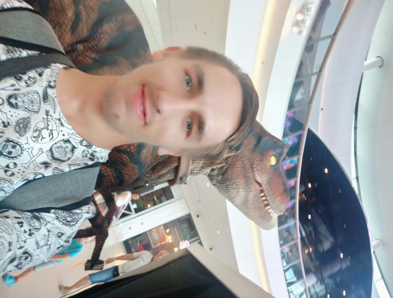 Młody mężczyzna pozuje do selfie z modelem dinozaura w jasnym, nowoczesnym centrum handlowym. Nietypowa pamiątka z wycieczki.