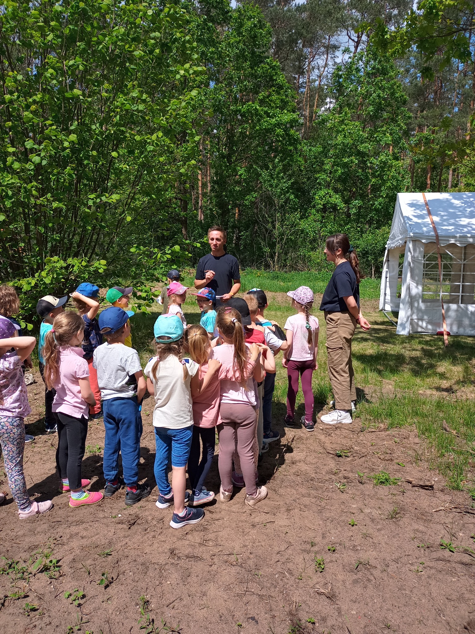 Grupa dzieci w różnokolorowych czapkach słucha dwóch dorosłych w lesie, w tle biały namiot.
