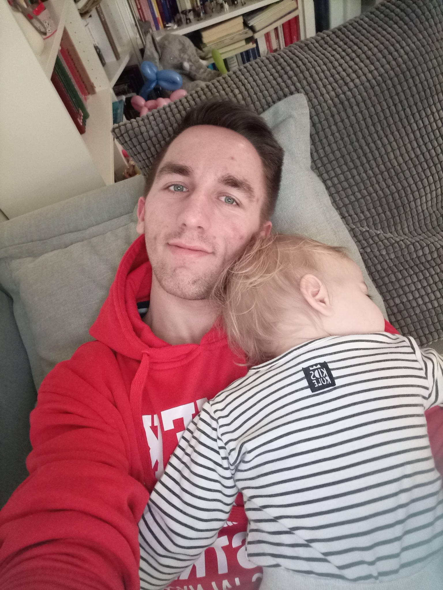 Mężczyzna w czerwonej bluzie z kapturem obejmuje śpiące dziecko w pasiastym ubranku na szarej kanapie w salonie z regałem z książkami w tle.