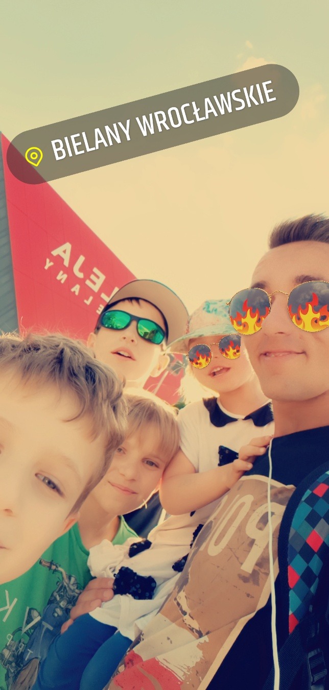 Grupa dzieci i młodych dorosłych pozuje do selfie na tle napisu 'Bielany Wrocławskie', wszyscy mają na sobie okulary przeciwsłoneczne, a niektórzy z nich mają nałożone filtry graficzne z płomieniami.