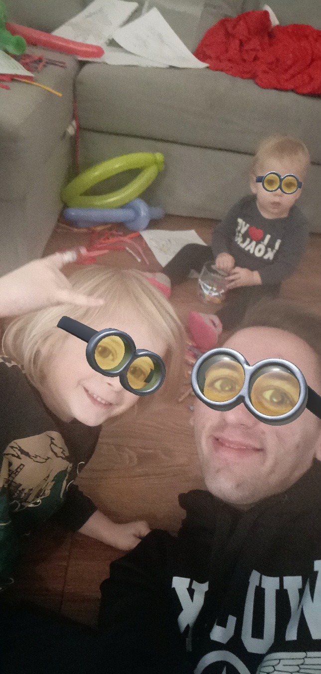 Zdjęcie zrobione z bliska przedstawia ojca z dwójką dzieci leżących na podłodze w salonie, wszyscy mają nałożony filtr z okularami Minionków, w tle widoczne balony i zabawki.