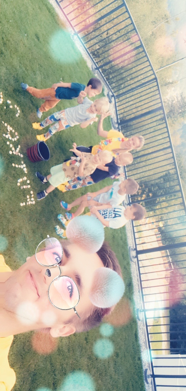 Zdjęcie z filtrem przedstawiające grupę dzieci i opiekuna na trawie, z napisem 'Happy' ułożonym z białych przedmiotów, obok stoi wiaderko.