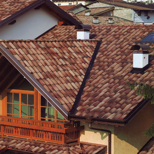 Widok z góry na dach pokryty włoską dachówką ceramiczną w odcieniach terakoty, z widocznym kominem i fragmentem balkonu z drewnianą balustradą. W tle inne dachy i budynki.