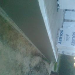 USŁUGI OGÓLNOBUDOWLANE KEN-BUD MACIEJ KĘDZIERSKI - Świeżo nałożona warstwa tynku na elewacji, widoczne białe narożniki i palety z bloczkami Solbet w tle.