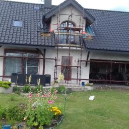 USŁUGI OGÓLNOBUDOWLANE KEN-BUD MACIEJ KĘDZIERSKI - Dom jednorodzinny w trakcie prac elewacyjnych, z rusztowaniami, zakrytymi oknami folią, widoczne kwiaty w ogrodzie i osoba na trawniku.