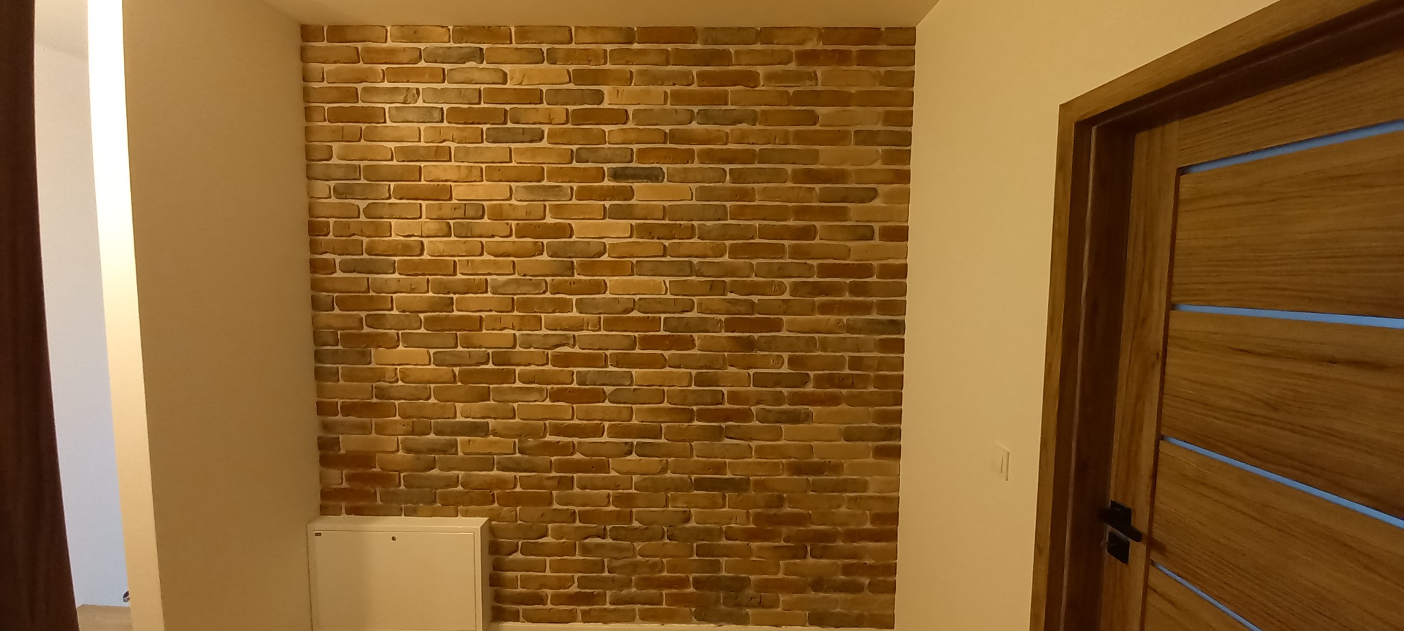 Ściana wewnętrzna wyłożona dekoracyjną cegłą w odcieniach brązu i beżu, z widocznym fragmentem drewnianych drzwi z poziomymi przeszkleniami i białą szafką przy podłodze.