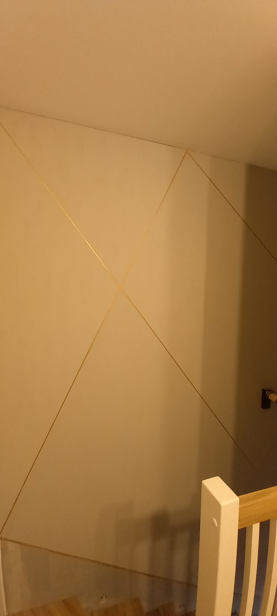 Beżowa ściana z geometrycznym wzorem z cienkich, złotych linii, widoczny fragment schodów i listwa przypodłogowa