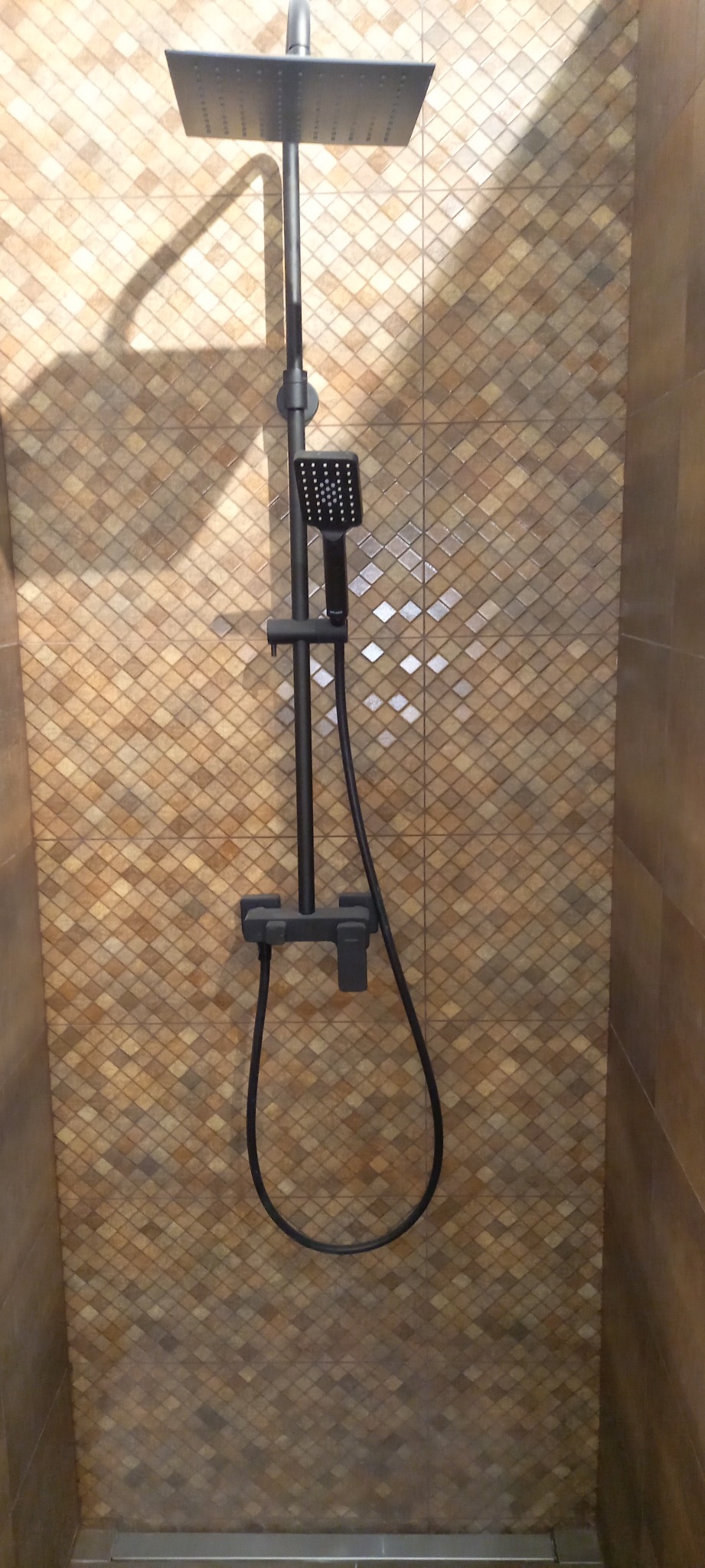 Czarny zestaw natryskowy z deszczownicą i słuchawką na tle mozaiki w odcieniach brązu i beżu z połyskującymi akcentami.