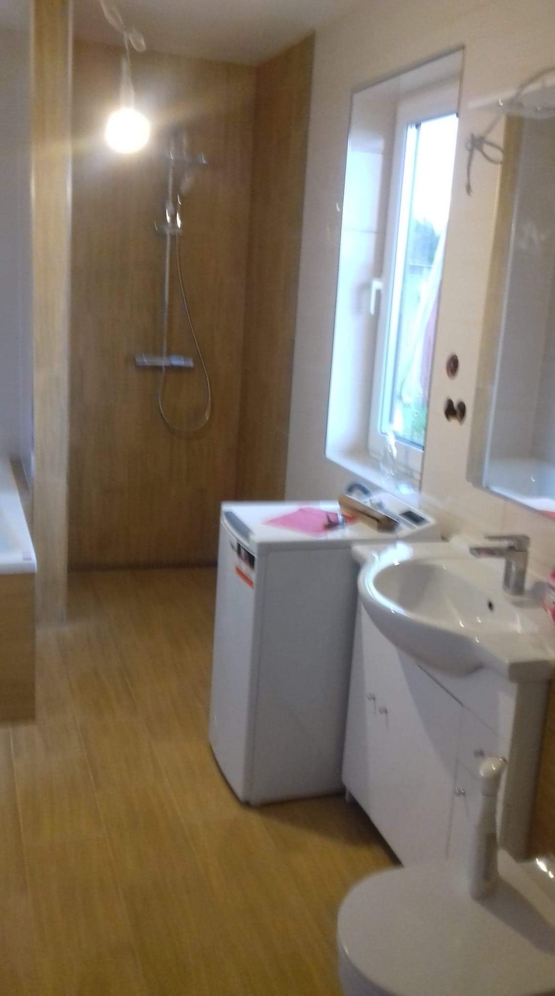 Wykończona łazienka z kabiną prysznicową, umywalką z szafką, pralką i drewnopodobną glazurą na ścianach i podłodze.