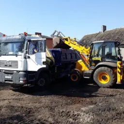 Żółto-czarna koparko-ładowarka JCB ładuje urobek do białej ciężarówki MAN na placu budowy z widocznym budynkiem w tle.