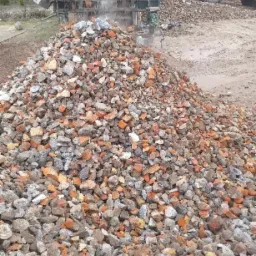 Góra gruzu ceglanego i betonowego pod kruszarką, z widoczną koparką w tle.