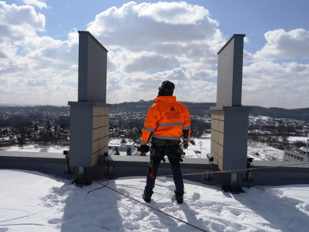 Pracownik w pomarańczowej kurtce stoi na zasypanym śniegiem dachu, między dwoma pionowymi elementami wentylacyjnymi, z widokiem na miasto w tle.