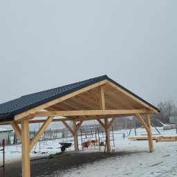 Art-Dach - Wznoszona drewniana konstrukcja z czarnym dachem z blachodachówki modułowej, widoczna w zimowej scenerii z ośnieżonym podłożem i mglistym tłem, na placu budowy z widocznymi narzędziami...