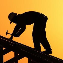 Art-Dach - Sylwetka dekarza w kasku, pracującego młotkiem na drewnianej konstrukcji dachu na tle pomarańczowego nieba o zachodzie słońca.