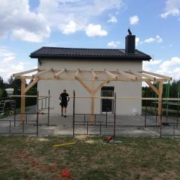 Art-Dach - Montaż drewnianej konstrukcji zadaszenia tarasu przy domu jednorodzinnym, widoczny pracownik w oddali oraz narzędzia na trawniku.