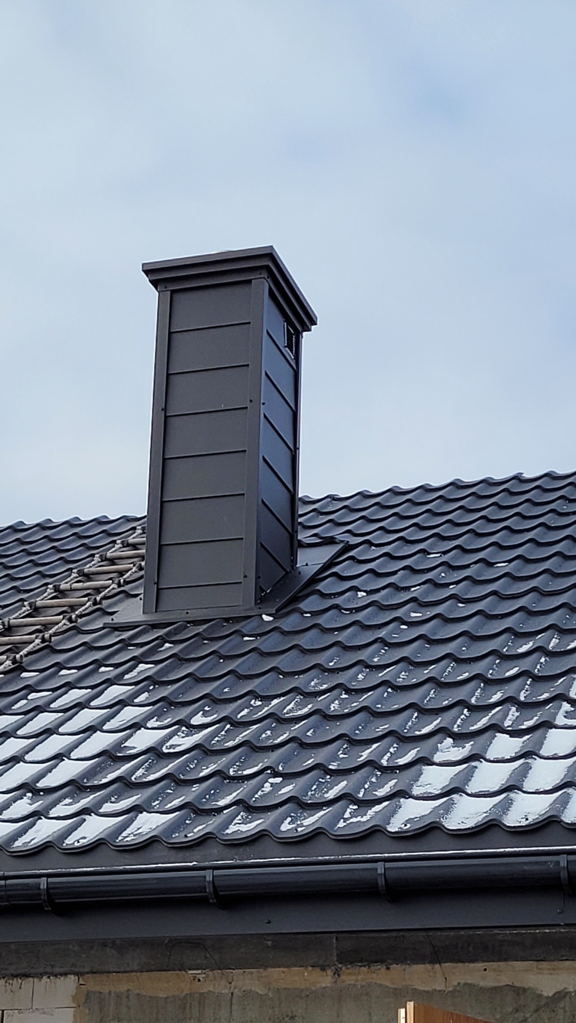 Nowy, grafitowy komin wentylacyjny z blachy na dachu pokrytym dachówką ceramiczną z resztkami śniegu, widoczne orynnowanie.