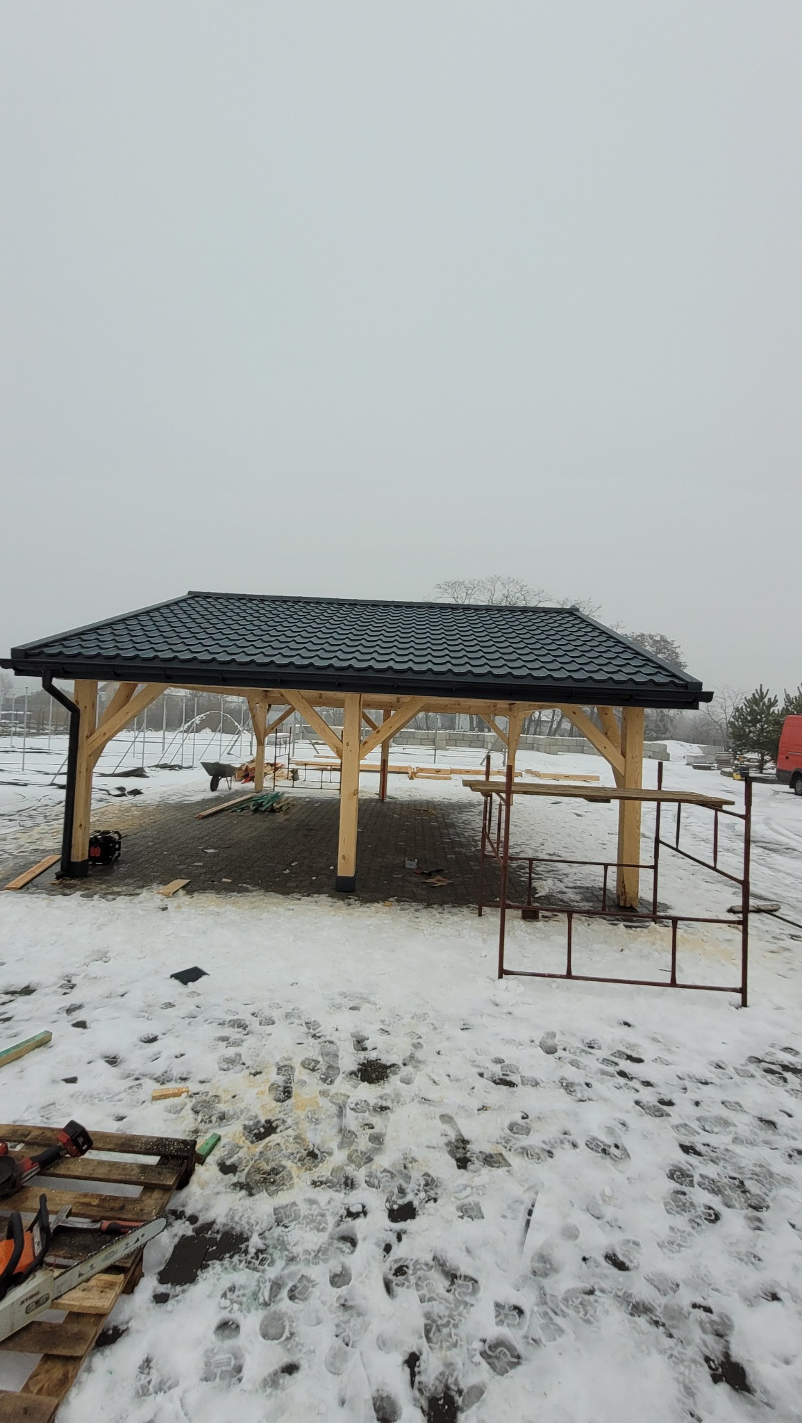Konstrukcja drewnianej altany z czarnym, metalowym pokryciem dachowym w trakcie montażu, otoczenie pokryte śniegiem, widoczne narzędzia i rusztowania.