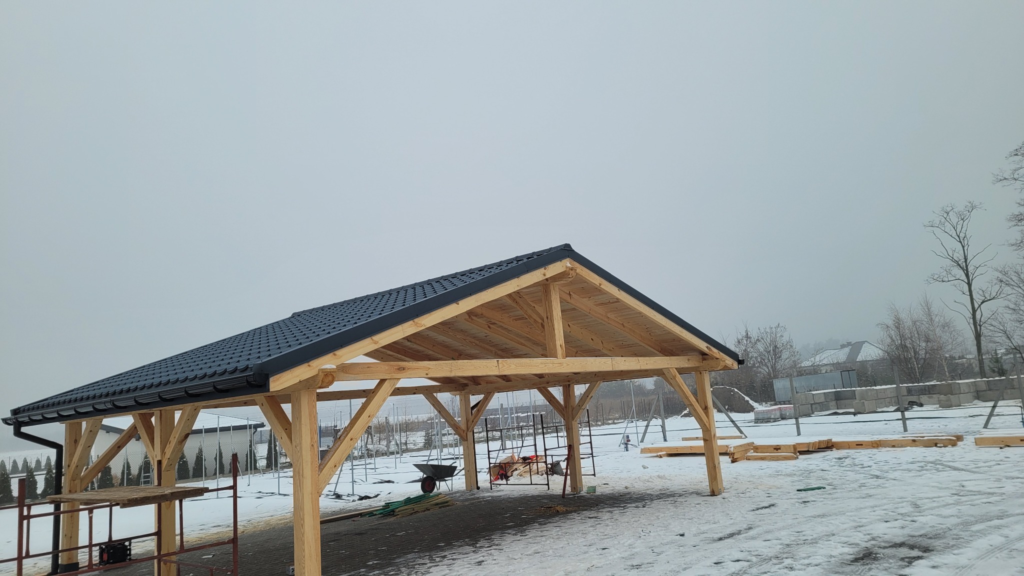 Wznoszona drewniana konstrukcja z czarnym dachem z blachodachówki modułowej, widoczna w zimowej scenerii z ośnieżonym podłożem i mglistym tłem, na placu budowy z widocznymi narzędziami...
