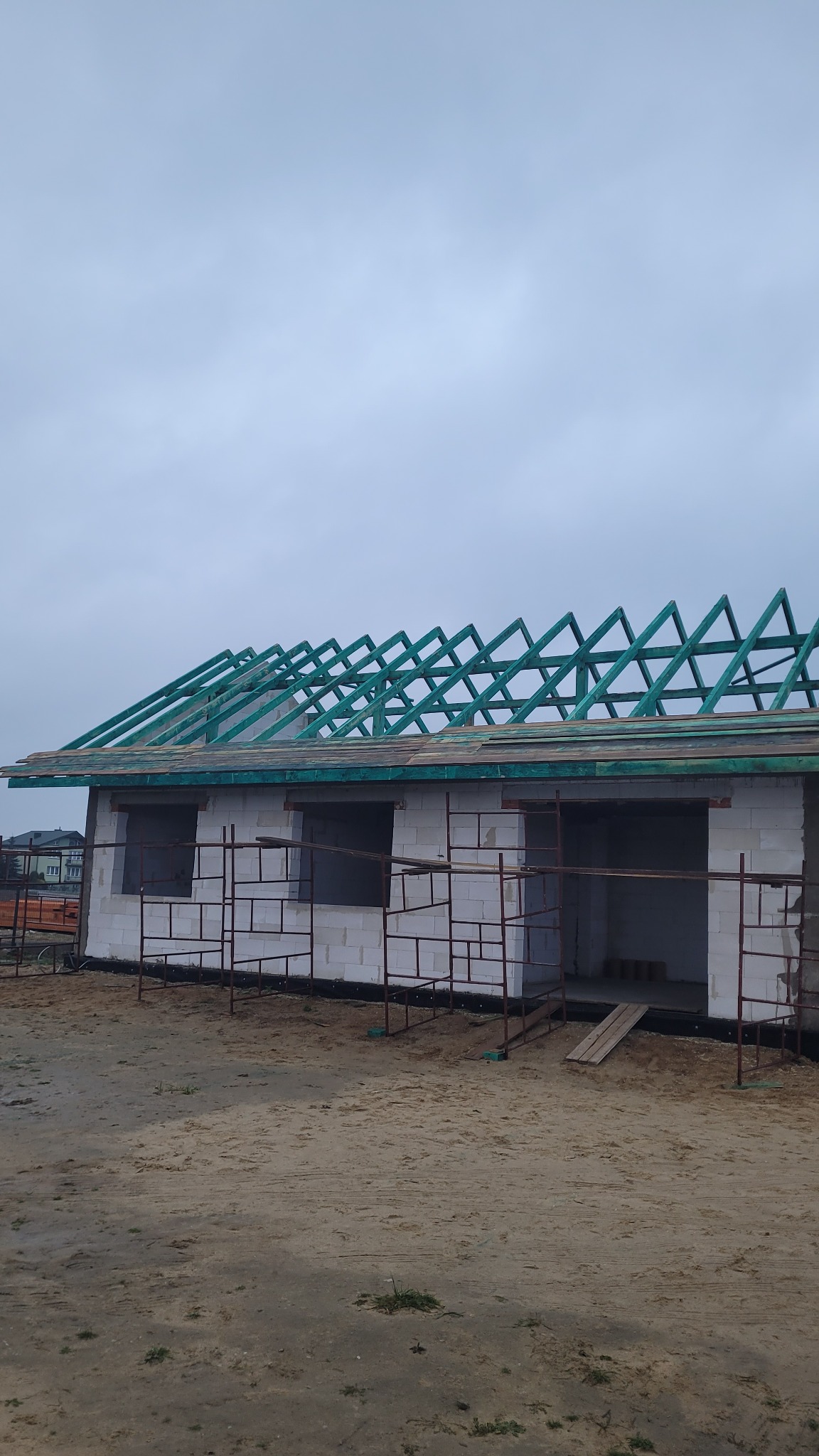 Niedokończony budynek z pustaków, z widoczną konstrukcją więźby dachowej z zielonego drewna i rusztowaniami, na piaszczystym terenie w pochmurny dzień.