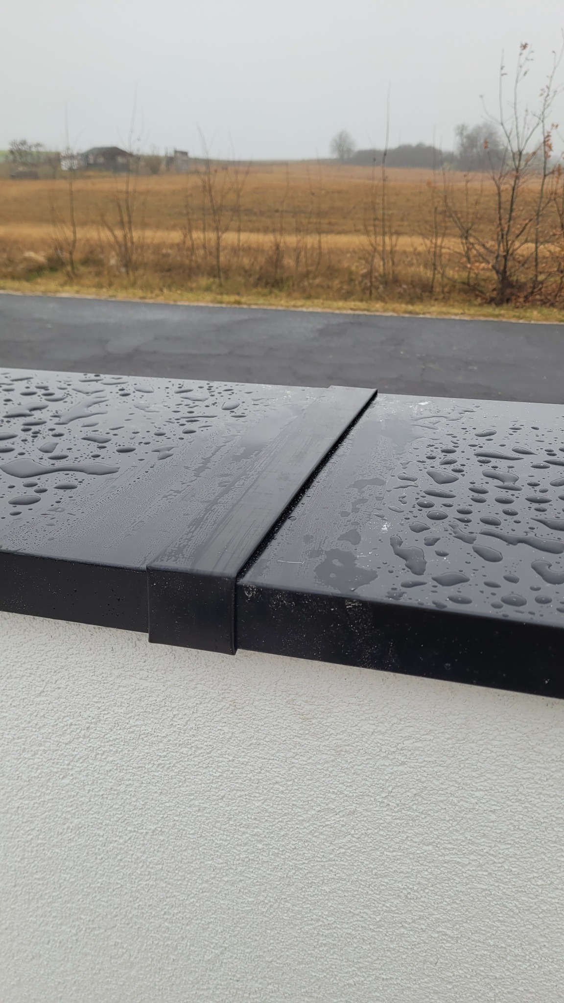 Metalowa obróbka blacharska na krawędzi dachu, pokryta kroplami deszczu, widok na szare pole i drzewa w tle.