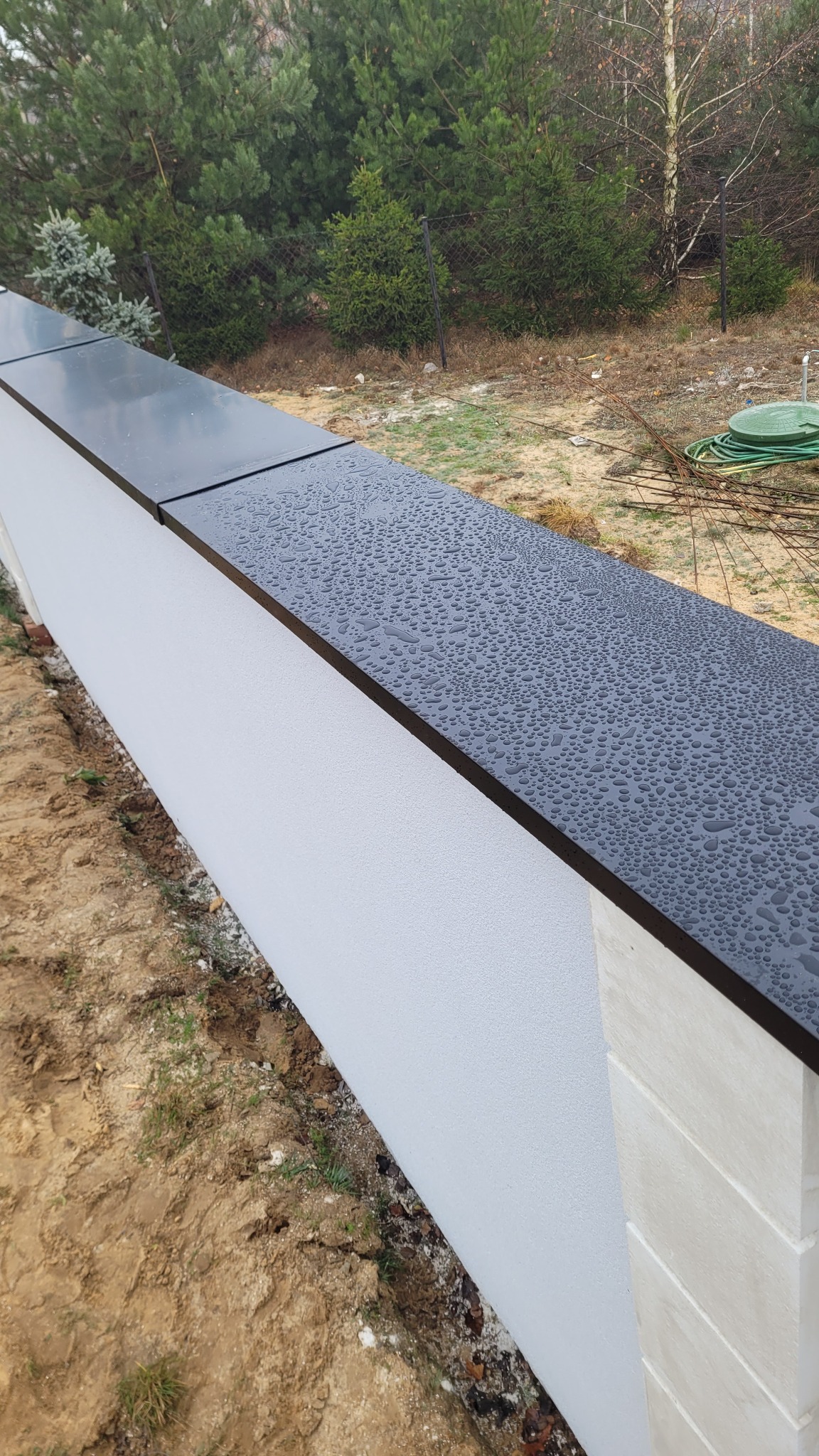 Nowoczesne, płaskie obróbki blacharskie na murku ogrodzeniowym, pokryte kroplami deszczu, z widoczną strukturą tynku i elementami dekoracyjnymi słupka.