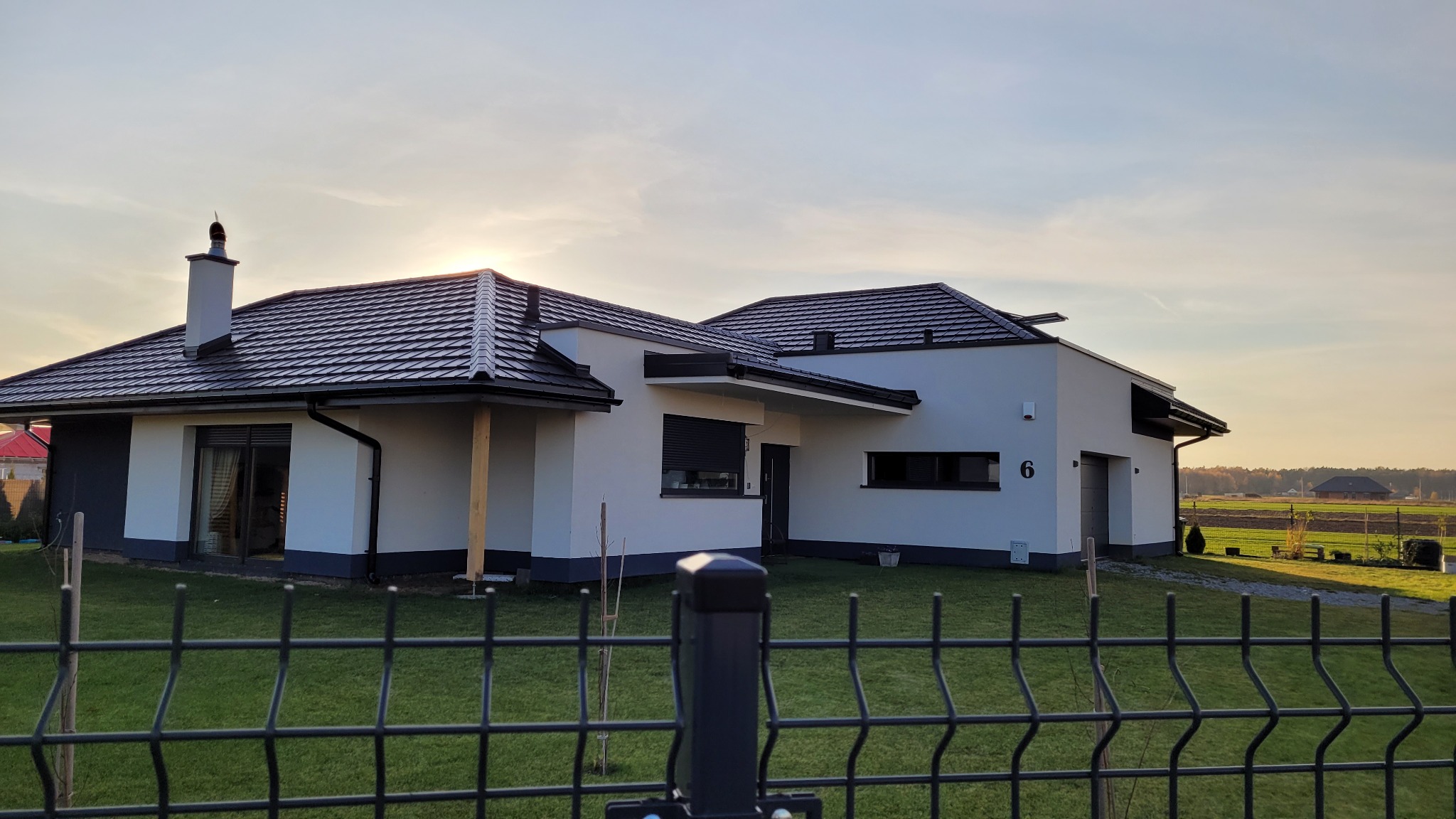 Nowoczesny dom jednorodzinny z ciemną dachówką ceramiczną, widok zza metalowego ogrodzenia, zachodzące słońce.