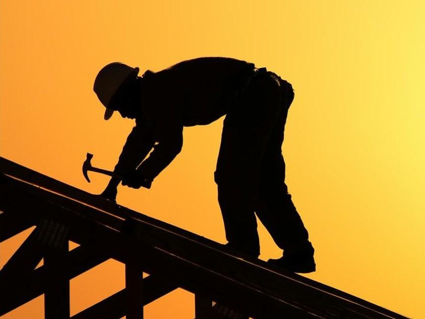 Sylwetka dekarza w kasku, pracującego młotkiem na drewnianej konstrukcji dachu na tle pomarańczowego nieba o zachodzie słońca.