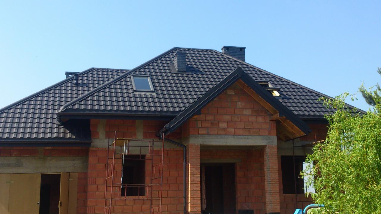 Dom w trakcie budowy z nowym, ciemnym dachem z dachówki ceramicznej, widoczne okno dachowe i komin, surowe ściany z czerwonej cegły, rusztowanie przy oknie.