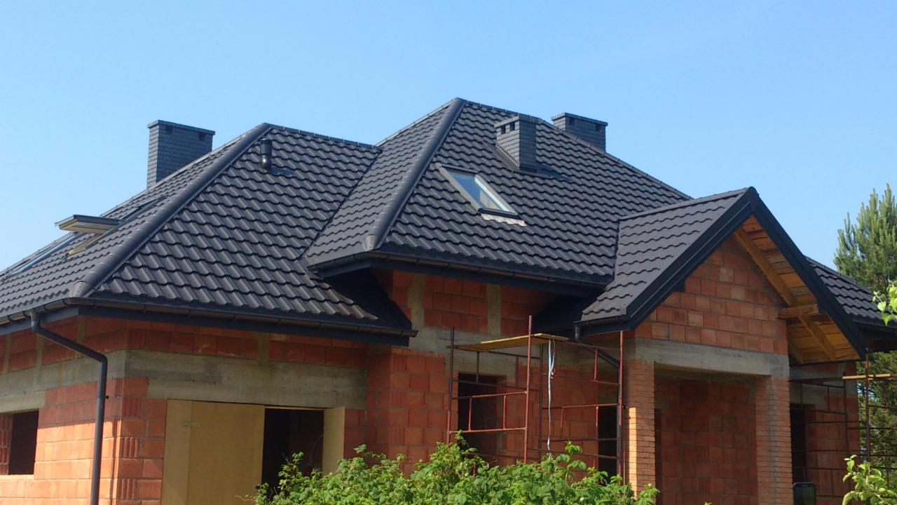 Dom w stanie surowym otwartym z nowym, ciemnym dachem z dachówki ceramicznej, widoczne rynny i okno dachowe, rusztowania przy elewacji.