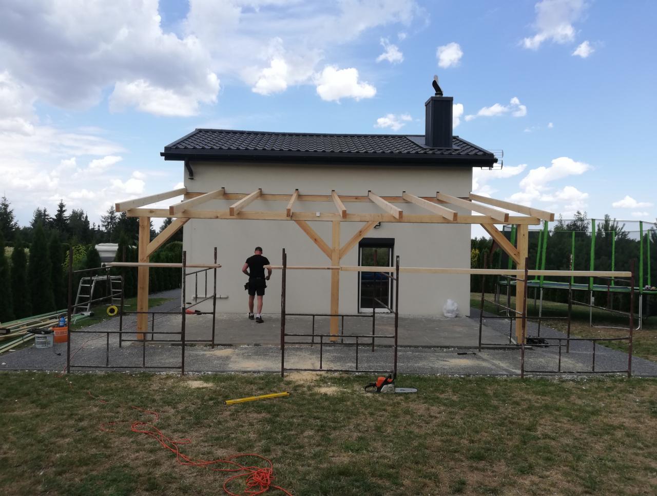 Montaż drewnianej konstrukcji zadaszenia tarasu przy domu jednorodzinnym, widoczny pracownik w oddali oraz narzędzia na trawniku.