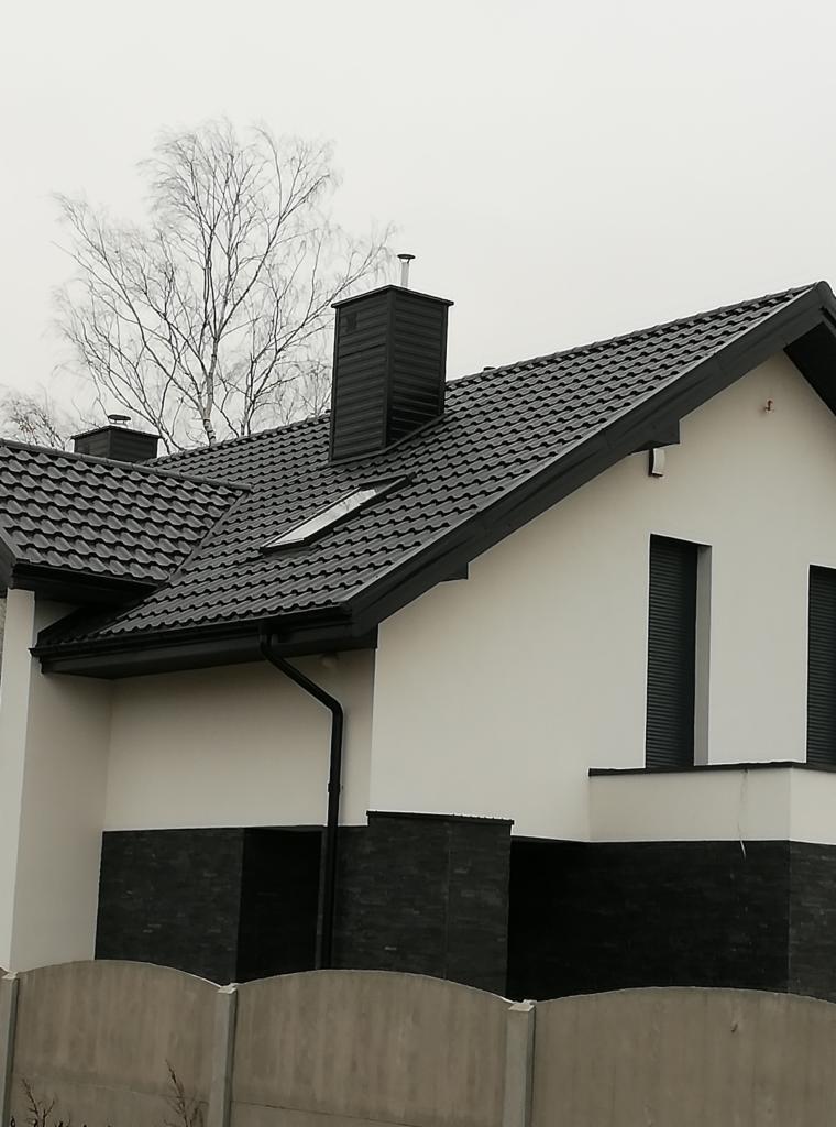 Nowoczesny dom jednorodzinny z ciemnoszarą dachówką, widoczne okno dachowe, komin i rynna spustowa, biała elewacja z ciemnym cokołem z kamienia, szara betonowa podmurówka.