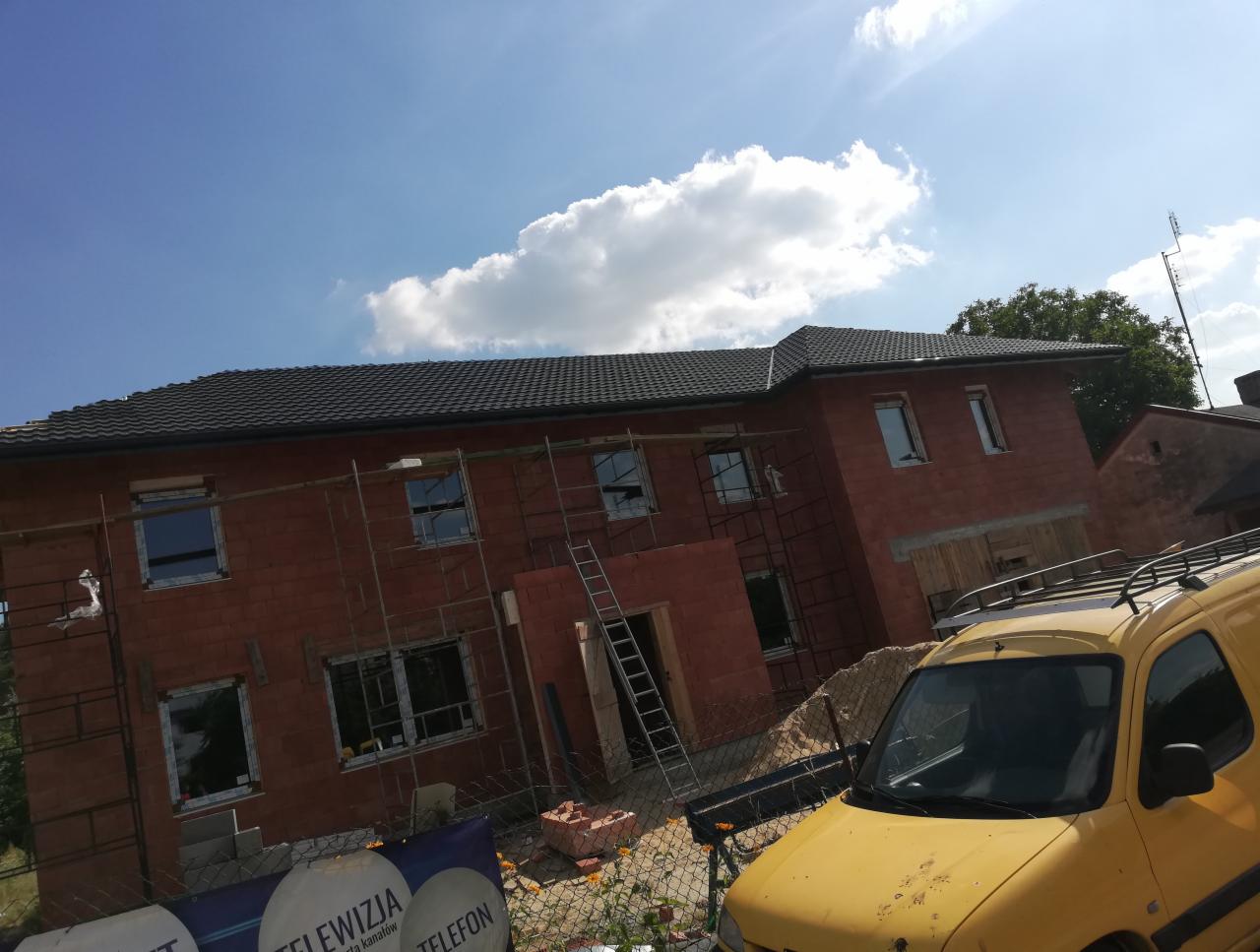 Budynek w trakcie budowy z nowym, ciemnoszarym dachem, widoczne rusztowania i żółty samochód dostawczy na pierwszym planie, błękitne niebo z chmurami w tle.