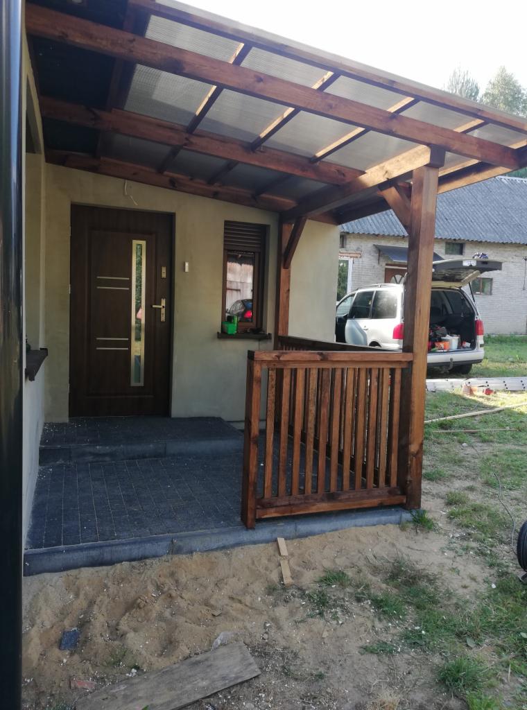 Drewniana konstrukcja z przezroczystym zadaszeniem nad wejściem do domu, z fragmentem drewnianej balustrady, widok na zaparkowany samochód i budynek gospodarczy w tle.