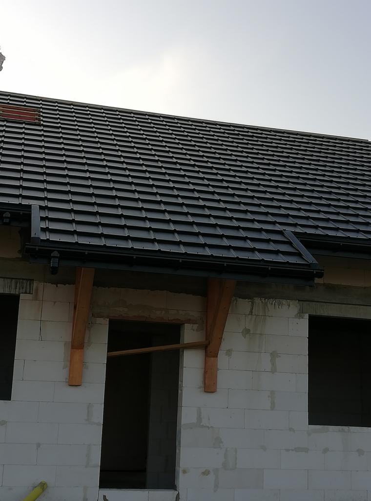 Nowy, czarny dach z rynnami na surowym budynku z białego bloczka, widoczne drewniane wsporniki.