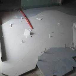 Układanie jasnych, kwadratowych płytek podłogowych z użyciem krzyżyków dystansowych, w tle fragment ciemniejszych, heksagonalnych płytek oraz poziomica.