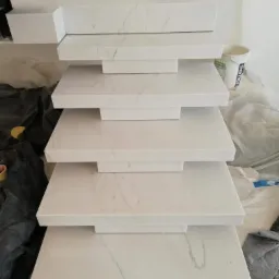 Stos wyciętych i obrobionych elementów z białego konglomeratu z szarymi żyłami, ułożonych jeden na drugim w warsztacie, prawdopodobnie przygotowanych do montażu jako parapety.