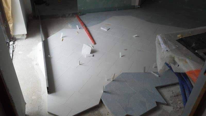 Układanie jasnych, kwadratowych płytek podłogowych z użyciem krzyżyków dystansowych, w tle fragment ciemniejszych, heksagonalnych płytek oraz poziomica.