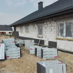 Dom w trakcie ocieplania, widoczne bloki styropianu Swissopor Lambda, częściowo przyklejone do ściany z białego bloczka, okna zabezpieczone folią, dach pokryty szarą dachówką.