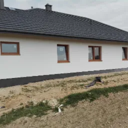 Świeżo otynkowana elewacja parterowego domu z brązowymi oknami, widok z boku na niezagospodarowany teren.