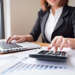 Kobieta w biurze, pracująca przy laptopie i kalkulatorze nad analizą wykres&oacute;w finansowych.