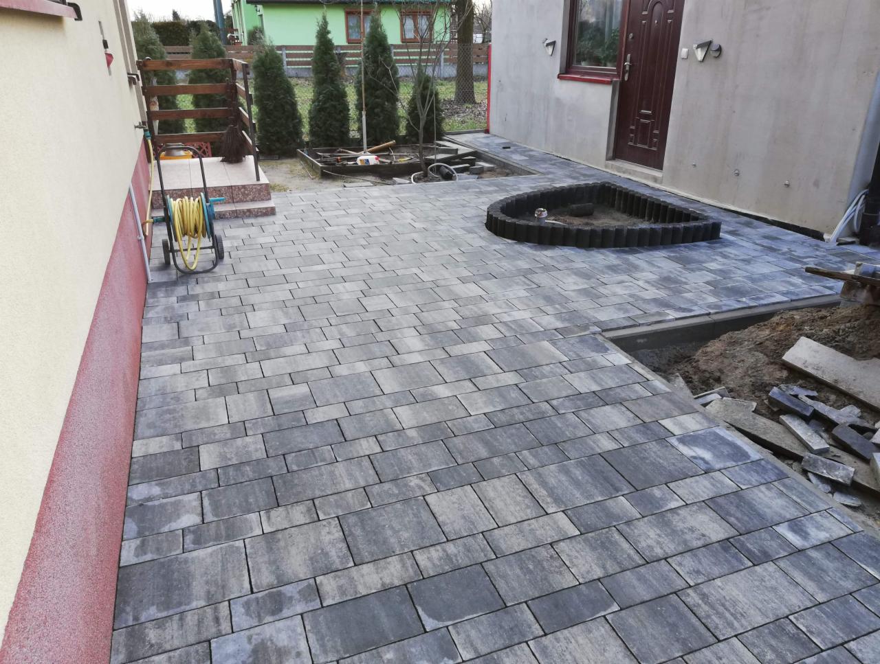 Świeżo ułożona, szara kostka brukowa na podjeździe, z fragmentem rabaty w kształcie łzy, widoczne narzędzia i pozostałości materiałów budowlanych.